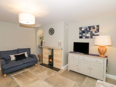 A living room with a blue sofa a small fireplace a wall clock a cabinet with a television and a lamp at Dazie Cottage in Broadway