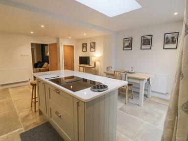 A kitchen with an island with stove and stools next to a dining table with chairs and framed pictures on the wall at Dazie Cottage in Broadway
