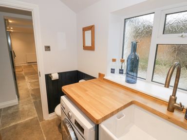 A utility room with a washing machine wooden countertop a sink and glass bottles on the windowsill at Dazie Cottage Broadway