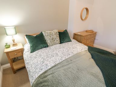 A bedroom with a bed green cushions a bedside table with a lamp and a dresser with a round mirror at Dazie Cottage in Broadway