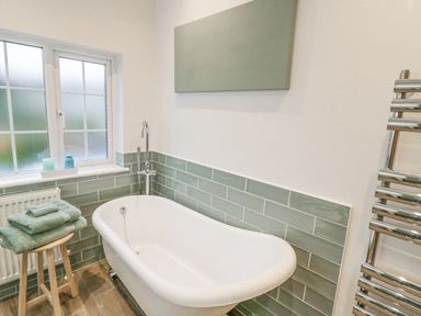 A bathroom with a white freestanding bathtub green tiles a wooden stool with folded towels a frosted window and a chrome towel rail at Dazie Cottage in Broadway