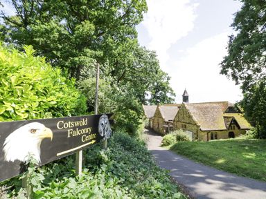 A pathway leading to stone buildings with a sign for Cotswold Falconry Centre surrounded by trees and bushes at Dazie Cottage in Broadway