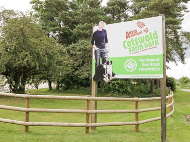 A wooden fenced area with a sign for Adam Hensons Cotswold Farm Park surrounded by trees
