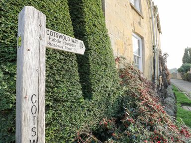 A wooden signpost for Cotswold Way public footpath next to a hedge and stone building at Dazie Cottage in Broadway