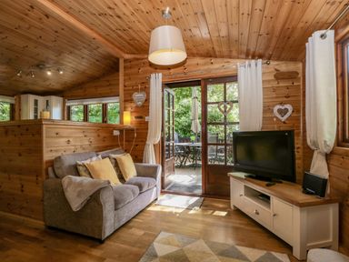 A living room with a couch, cushions, a television on a stand, French doors leading to an outdoor patio with a table and chairs at Scafell Lodge Skiptory Howe 7, White Cross Bay Holiday Park near Windermere