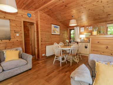 A wooden dining area with a round table and white chairs next to a living room with gray sofas and yellow cushions at Scafell Lodge Skiptory Howe 7 White Cross Bay Holiday Park near Windermere