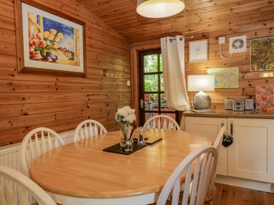 A dining area with a wooden table and white chairs next to a wooden wall with a framed painting and a cabinet with a lamp and kitchen items at Scafell Lodge Skiptory Howe 7 White Cross Bay Holiday Park near Windermere
