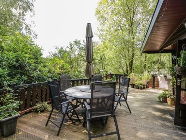 An outdoor wooden deck with a round table and six chairs around it and plants around the railing at Scafell Lodge in Skiptory Howe 7 White Cross Bay Holiday Park near Windermere