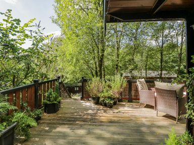 A wooden deck with potted plants and wicker chairs with cushions surrounded by trees at Scafell Lodge in Skiptory Howe 7 White Cross Bay Holiday Park near Windermere