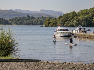 Two people paddleboarding on a lake near boats and a dock with trees and mountains in the background at Scafell Lodge Skiptory Howe 7 White Cross Bay Holiday Park near Windermere