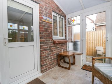 A small enclosed porch with a tiled floor two white doors a brick wall a wooden side table and a wicker chair with a cushion