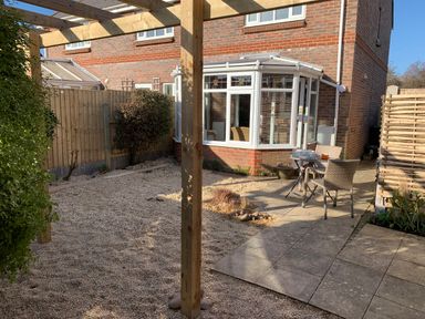 A backyard with gravel ground a wooden pergola a patio with a small table and two chairs and a brick house with a white conservatory