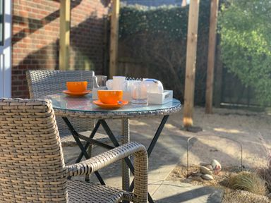 An outdoor patio with a round glass table two woven chairs orange cups and a white tray with teapots and cups at The Beach House in West Bay