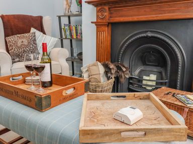 A room with an upholstered chair cushions wooden trays wine bottle and glasses by a fireplace at Anchor Cottage in Whitby