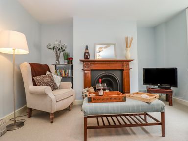 A sitting room with an armchair next to a floor lamp a coffee table with trays and wine glasses a fireplace a shelf with books and a television at Anchor Cottage in Whitby