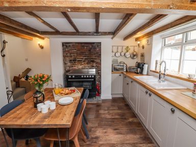 A kitchen with wooden beams on the ceiling a wooden dining table with chairs a brick wall with a stove and a counter with a sink at Anchor Cottage in Whitby