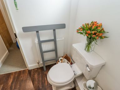 A bathroom with a white toilet flowers in a glass vase on the toilet tank a basket with toilet paper rolls and a grey towel rack against the wall at Anchor Cottage in Whitby