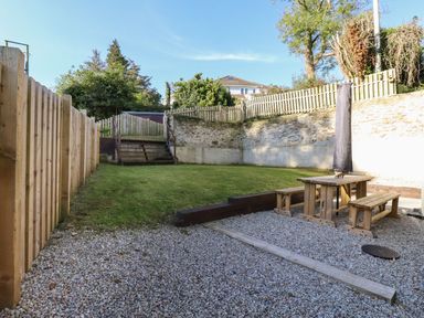 A backyard with a gravel seating area and wooden benches next to a grassy lawn and wooden fence at Brewers Cottage in Looe