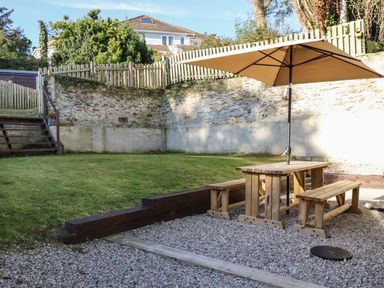 A garden area with a wooden picnic table and benches under a large umbrella on gravel next to a grassy lawn and stone wall at Brewers Cottage in Looe