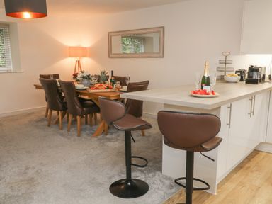 A dining area with a wooden table, six chairs, a mirror on the wall, and a kitchen counter with two bar stools and a bottle of champagne with glasses at Brewers Cottage in Looe