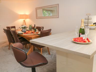 A dining area with a wooden table set with plates and croissants and a kitchen counter with champagne and strawberries at Brewers Cottage in Looe