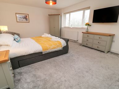 A bedroom with a bed, bedside table with lamp, wardrobe, dresser with flowers and wall-mounted TV at Brewers Cottage in Looe