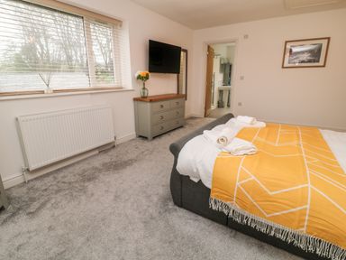 A bedroom with a bed covered with a yellow patterned throw and rolled towels a dresser with a vase of flowers a wall mounted television and an open door showing a bathroom at Brewers Cottage in Looe