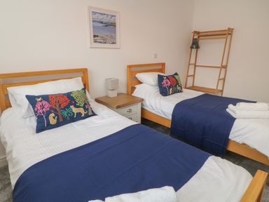 A bedroom with two single beds with blue blankets and decorative pillows a bedside table with a lamp a wooden clothes rack and a framed picture on the wall at Brewers Cottage in Looe