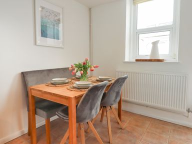 A dining area with a wooden table set with plates and a vase of tulips two gray chairs and a bench under a window at Quayside Cottage in Teignmouth