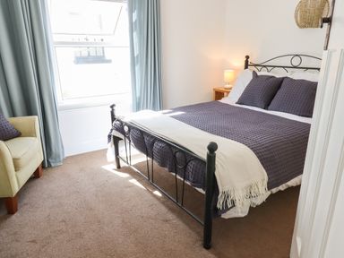 A bedroom with a black metal bed frame with purple bedding and white throw beside a window with blue curtains and a yellow armchair at Quayside Cottage in Teignmouth