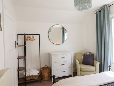 A bedroom corner with a white dresser under a round mirror next to a yellow armchair by a window with blue curtains at Quayside Cottage in Teignmouth