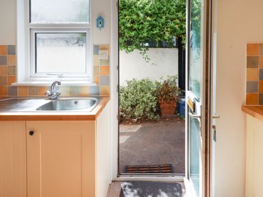 A kitchen sink and counter with tiled backsplash next to an open door leading outside to a garden area at Quayside Cottage in Teignmouth