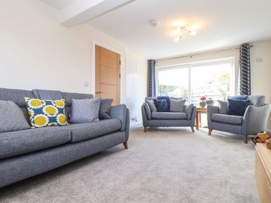 A living room with three grey sofas cushions a wooden door and large window with curtains at Pickney House in Killigarth near Polperro