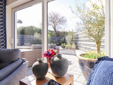 A seating area with vases and a flower vase on a wooden table next to large glass doors opening to a patio with outdoor furniture and plants at Pickney House in Killigarth near Polperro