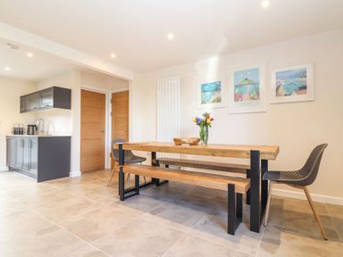 A dining area with a wooden table, benches, two black chairs, kitchen cabinets, and three framed pictures on the wall at Pickney House in Killigarth near Polperro