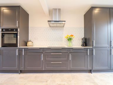 A kitchen with grey cabinets a built in oven a stove a glass vase with flowers and a tiled backsplash at Pickney House in Killigarth near Polperro