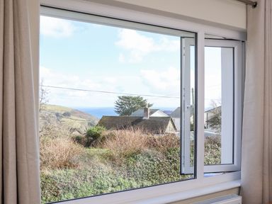 A view of rooftops shrubs hills and sea through an open window with beige curtains at Pickney House Killigarth near Polperro