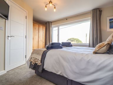 A bedroom with a bed, folded towels on the bed, a wooden wardrobe, a window with curtains and a door at Pickney House in Killigarth near Polperro