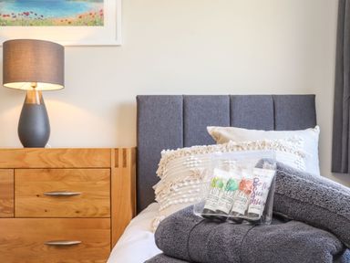 A bedroom with a wooden bedside table with a lamp next to a bed with gray headboard and gray towels on the bed at Pickney House Killigarth near Polperro