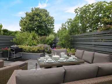 An outdoor patio with cushioned wicker seating and a table set with plates and cups next to plants and a barbecue at Pickney House in Killigarth near Polperro