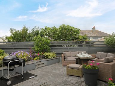An outdoor patio with a BBQ grill, wicker seating, a dining table set with dishes, and plants at Pickney House in Killigarth near Polperro
