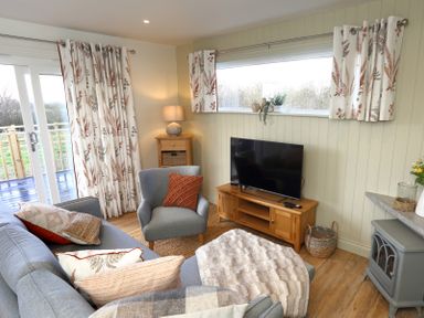 A living room with a gray sofa, an armchair, a wooden TV stand with a TV, a side table with a lamp, patterned curtains, and a small fireplace at The Swift in Wem