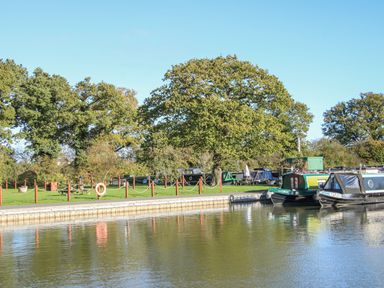 A canal with narrowboats moored along a grassy bank with large trees and a picnic table at The Swift in Wem