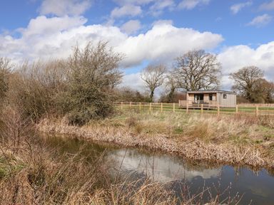 A small wooden cabin near a fenced grassy area beside a pond with trees and bushes under a cloudy sky at The Swift in Wem