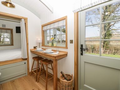 A small dining area with two wooden stools a wooden table a basket with firewood beside a door and a window with flowers at Acorn in Bleadon