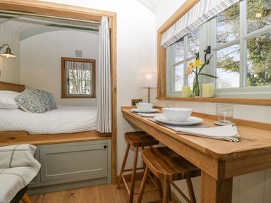 A small dining area with two wooden stools and table set with bowls and plates next to a bed with floral pillows at Acorn in Bleadon