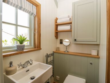 A small bathroom with a white sink a window with a plant on the sill shelves with towels and a closed cabinet at Acorn in Bleadon
