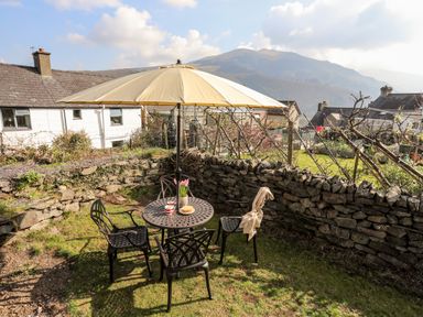 A garden seating area with a round metal table under a large umbrella and four chairs surrounded by a stone wall at 9 Ty Du Road in Llanberis