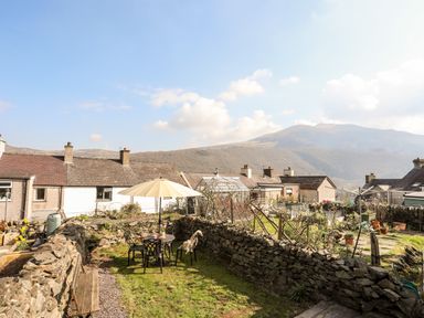 A garden with stone walls a table with chairs and an umbrella and houses and hills in the background at 9 Ty Du Road in Llanberis