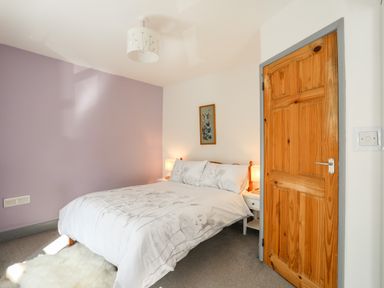 A bedroom with a double bed with white bedding and two bedside tables with lamps a wooden door and a purple accent wall at 9 Ty Du Road in Llanberis